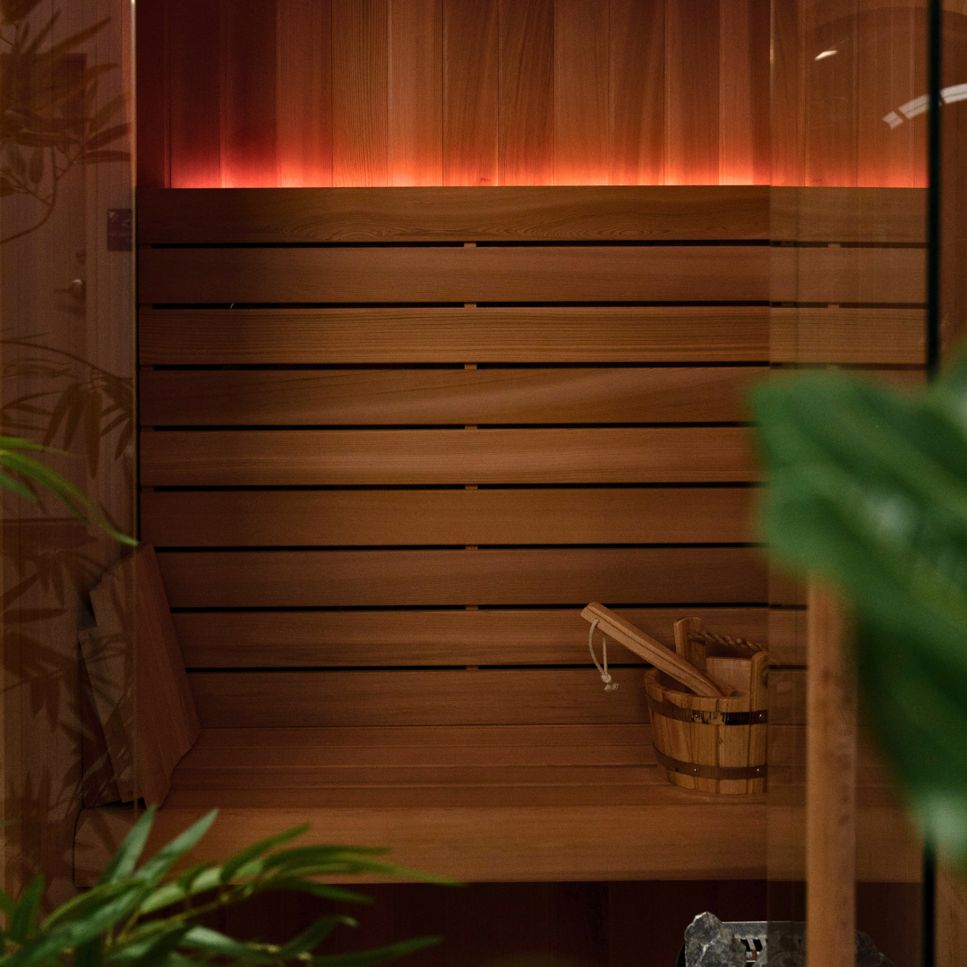 Wooden sauna with a bucket and wooden bench, surrounded by plants.
