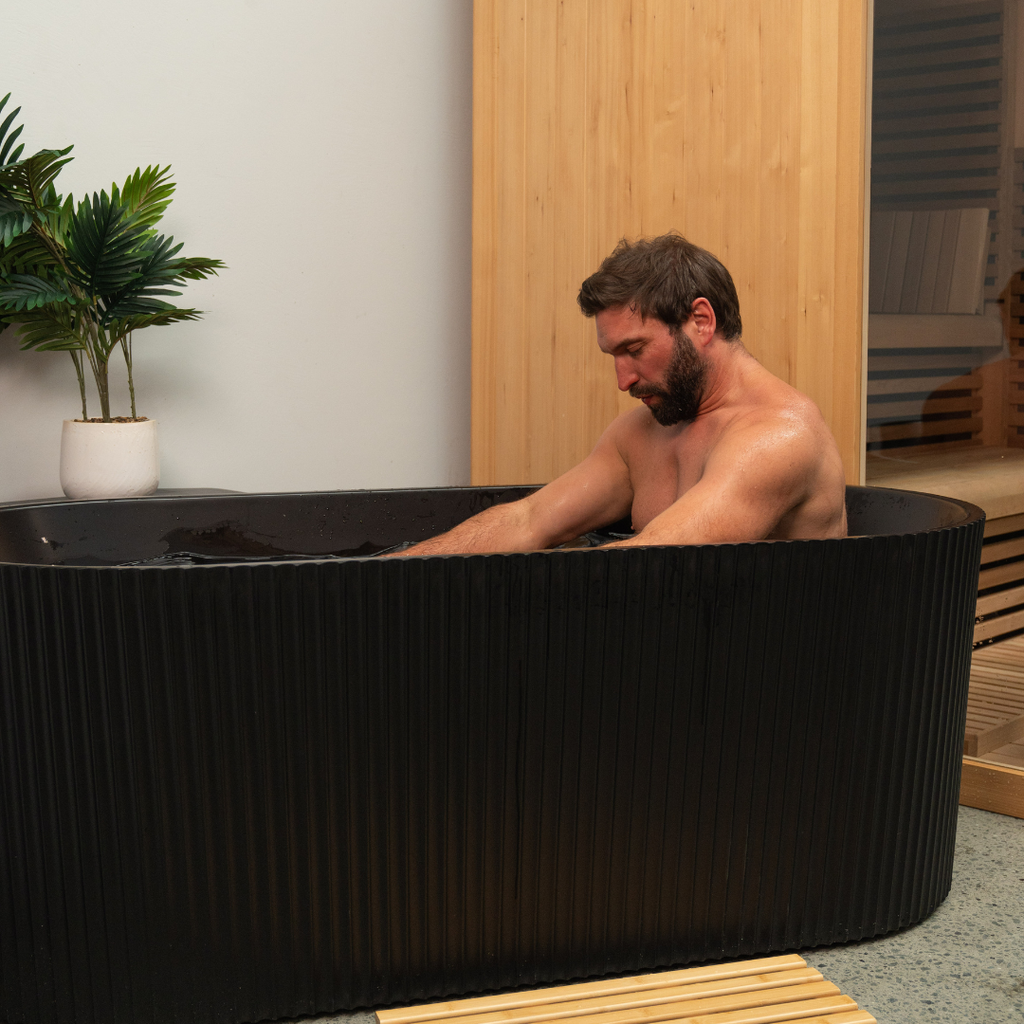 Man sitting in a black freestanding bathtub in a modern room with wooden walls and a plant.