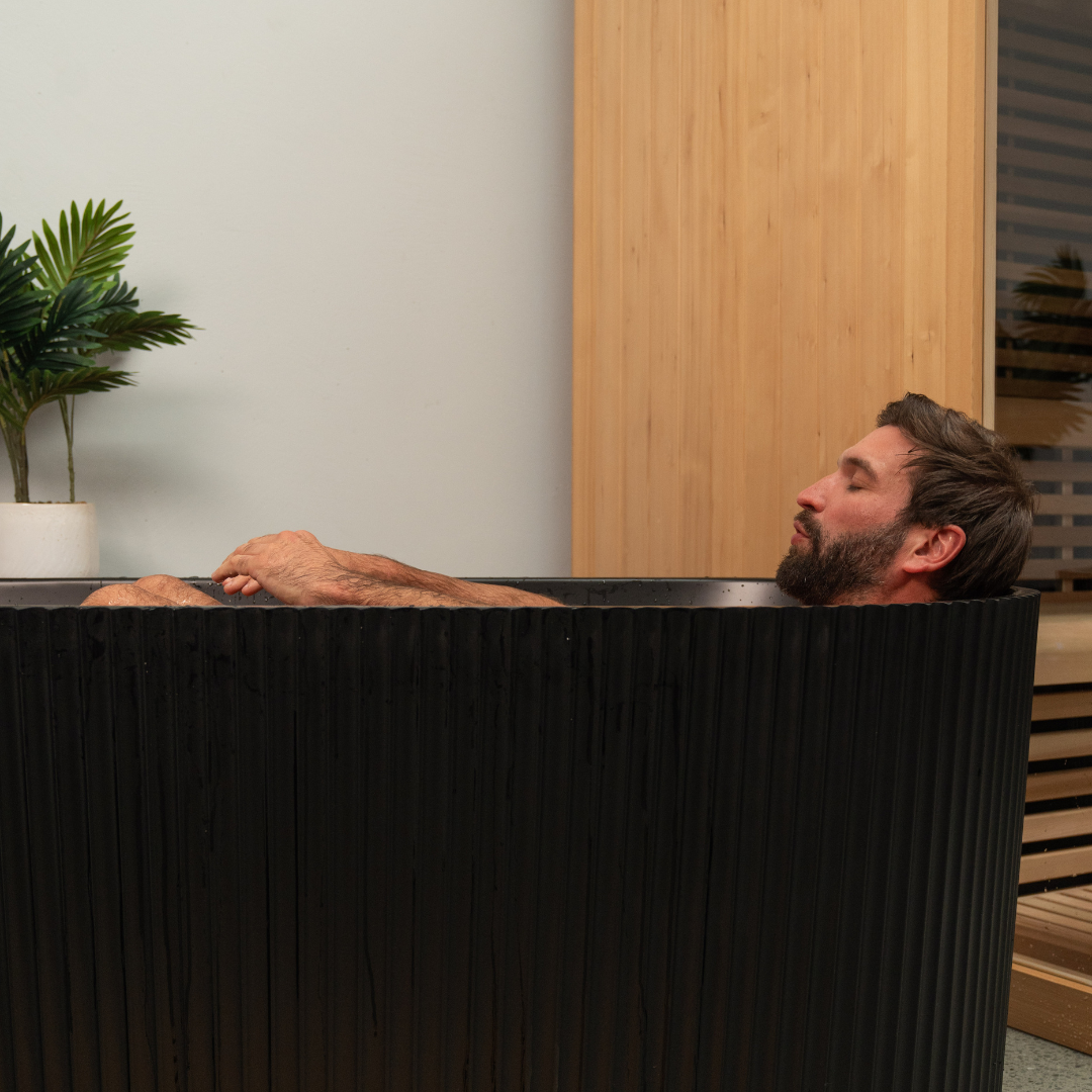 Man relaxing in a modern black bathtub with a plant and wooden panel in the background