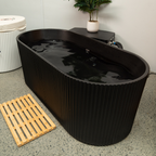 Black acrylic ice bath with water inside, placed on a concrete floor with a wooden mat and plant in the background.