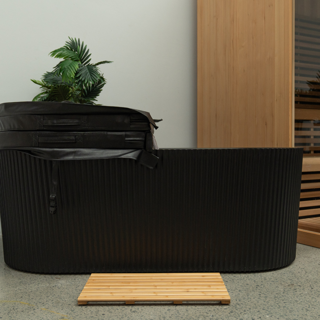Black outdoor ice bath with a wooden mat on a concrete floor, next to a wooden bench and plant.