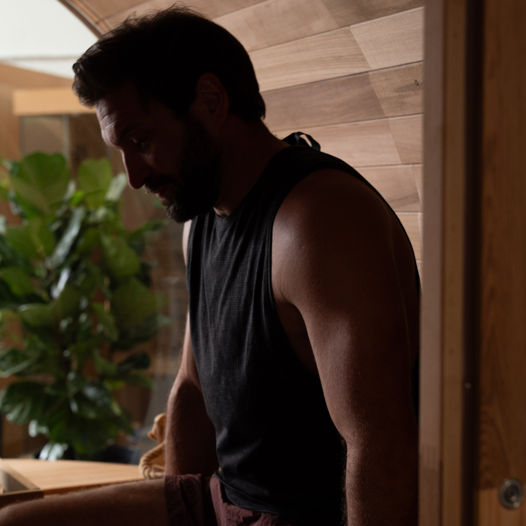 Man in a black tank top standing in a room with wooden walls and a plant.